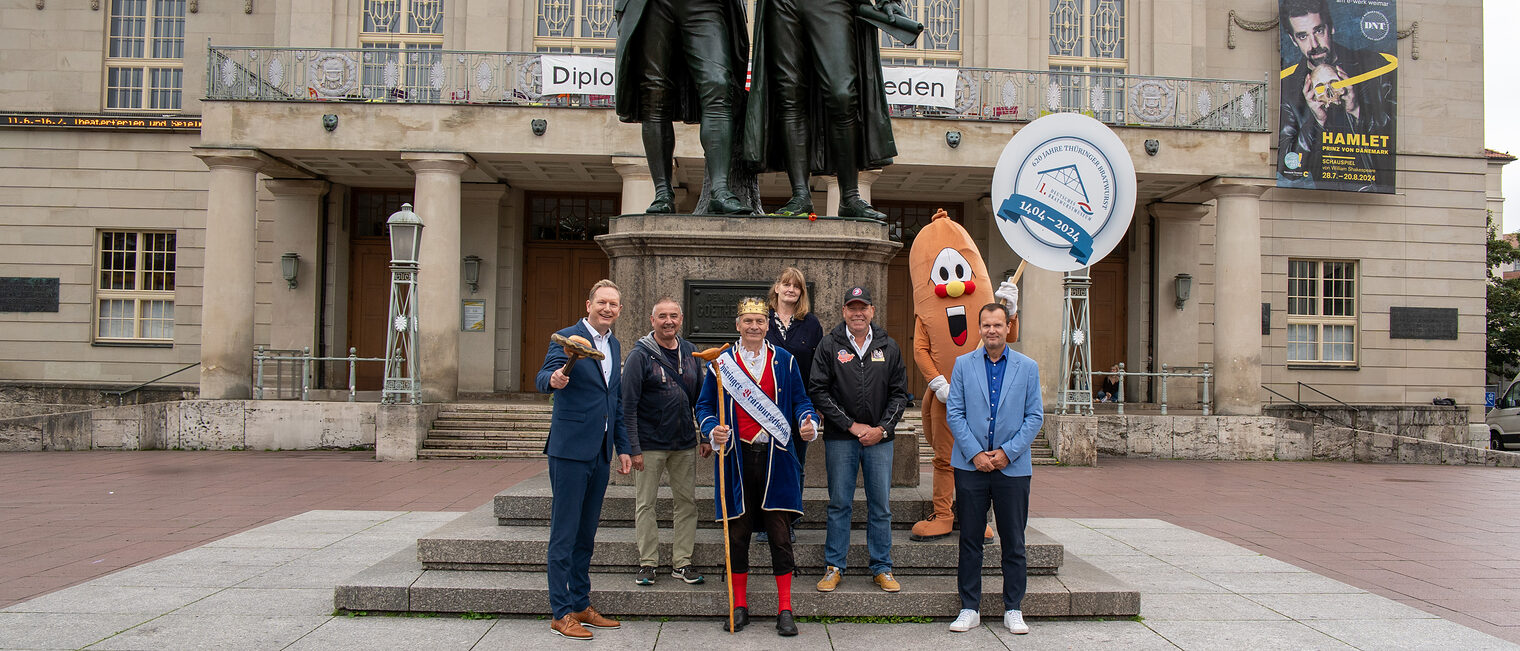 Die Initiatoren und Unterstützer des Wurst- und Spezialitätenmarktes vor dem Goethe- und Schiller-Denkmal in Weimar (v.l.n.r.): Tobias Hinz (Vorstandsvorsitzender des Vereins zur Förderung des Handwerks Thüringen e.V.), Frank Martin (Vorstandsmitglied des Vereins zur Förderung des Handwerks Thüringen e.V.), Thüringer Bratwurstkönig Norbert I, Helgard Anding (Geschäftsführerin des Landesinnungsverbandes des Fleischerhandwerks Thüringen e.V.), Thomas Mäuer (Geschäftsführer der Bratwurstmuseum Betriebs-GmbH), Christian Schwartze (Amtsleiter für Wirtschaft und Märkte Weimar) und Maskottchen „Wursti“.